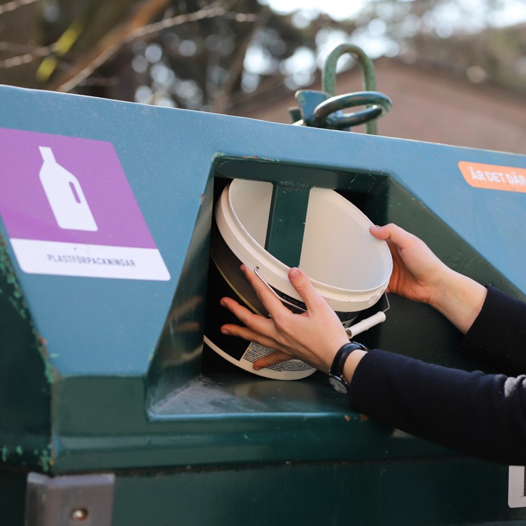 Närbild på händer som lägger en plastbunke i behållaren för plastförpackningar på en återvinningsstation.